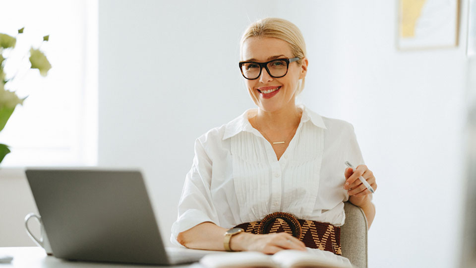 Frau mit Brille und in weißer Bluse sitzt vor einem Laptop und lächelt in die Kamera.